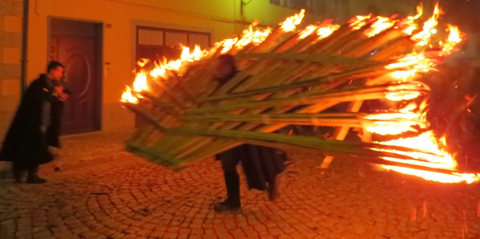 Portatore in mantello scuro con una grande 'ndoccia a ventaglio in fiamme su una strada lastricata di Agnone, di notte